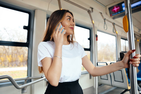 A young woman is engaged in a conversation on her mobile phone while holding onto a pole inside a tram. Sunlight filters through the windows, illuminating the interior space.の写真素材