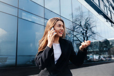 A young woman is standing outside a contemporary building, speaking enthusiastically on her smartphone while gesturing with her hands, clearly enjoying her conversation during daytime.の写真素材