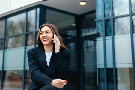 A young woman dressed in a black suit sits casually while talking on her phone outside a contemporary office building, smiling and enjoying the moment.の写真素材