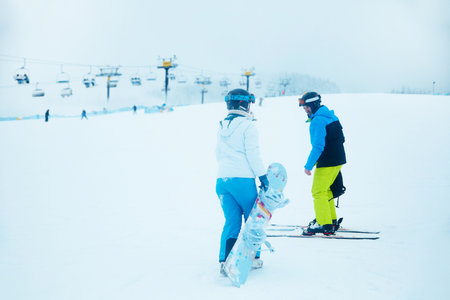 Two winter sports enthusiasts prepare for fun on the slopes as they gear up with skis and a snowboard at a busy ski resort on a chilly day.の写真素材
