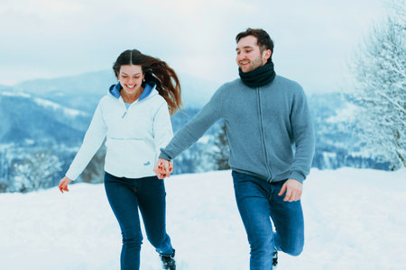A young couple walks hand in hand through the winter snow, laughing and enjoying the chilly air in a...