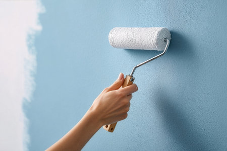 A person is holding a paint roller with a wooden handle. The roller is covered in white paint and is being used to apply the paint to a light blue wall.の素材