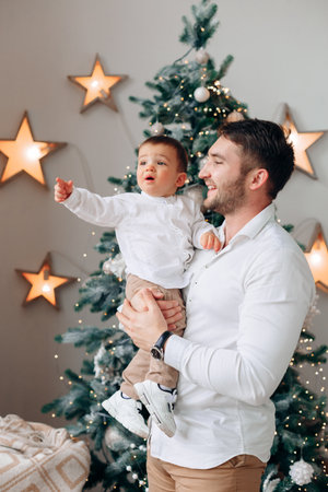 A father smiles and lifts his cheerful son in front of a beautifully decorated Christmas tree with glowing lights and star ornaments, embracing the festive spirit.の写真素材