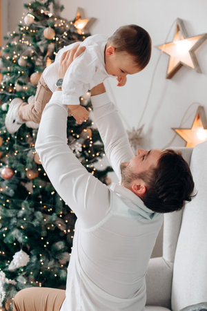 In a cozy room, a father lifts his smiling child high in the air. A beautifully decorated Christmas tree, full of lights and ornaments, sets a festive mood.の写真素材