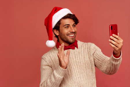 Smiling man wearing a Christmas hat and sweater is taking a selfie with his smartphone while waving cheerfully against a warm holiday-themed backdrop.の素材