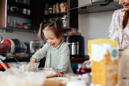 A young child smiles and stirs a bowl of cake batter in a busy kitchen. An adult stands nearby,...