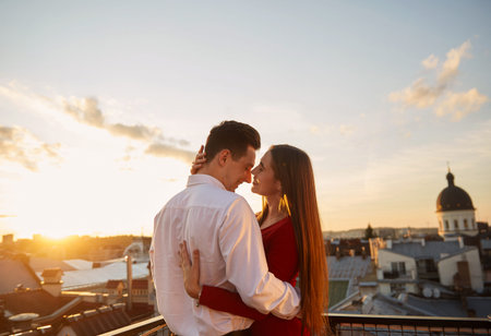 Two people stand close on a rooftop while watching the sunset over the city skyline during the...