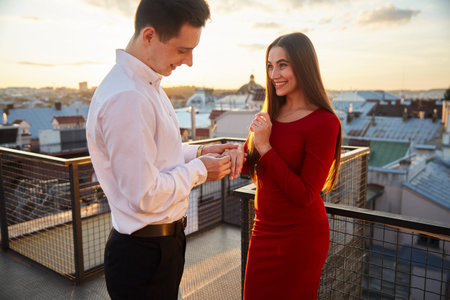 A man presents a ring to a woman while they stand on a rooftop overlooking the city at sunset.