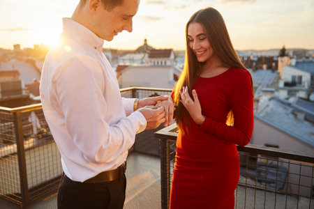 Man shows ring to woman during proposal on balcony. Sun sets behind them as they stand on a rooftop...