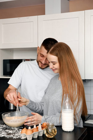 Two people are standing in a kitchen, mixing batter in a bowl. They smile at each other while...