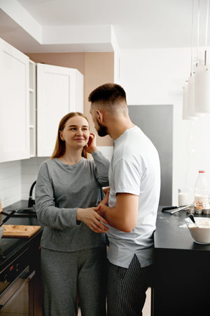 Man and woman stand close in a kitchen. They smile and touch each others faces. Cooking utensils are...