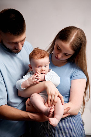 Two adults hold and look at their baby together in a studio. The baby seems curious and is sucking...