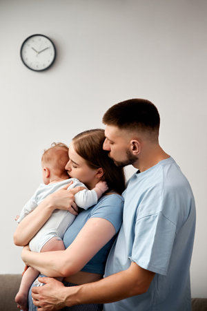 A couple holds their baby in a living room with a clock on the wall. They stand close to each other,...