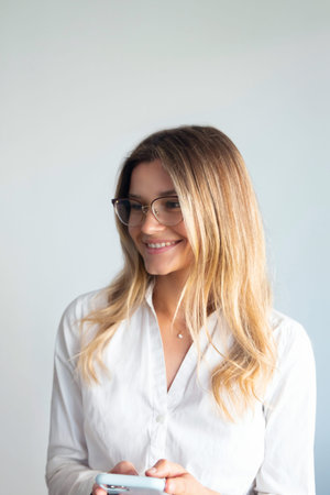 A woman stands in an office, smiling while looking at her phone. She wears glasses and is dressed in a white shirt.の写真素材