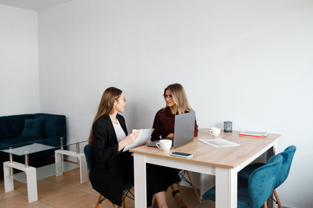 Two women sit in an office meeting. They review documents and talk about work at a table with a...