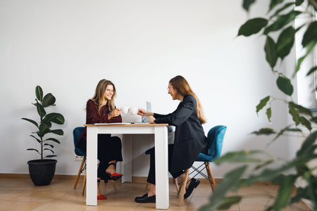 Two women are seated at a table in an office, engaging in conversation and enjoying drinks while...