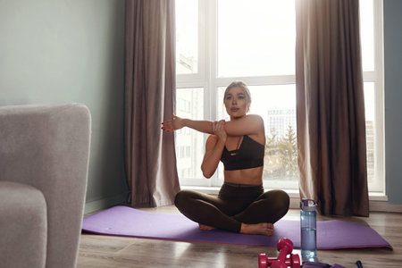 A woman sits on a yoga mat in her home and stretches her arm while preparing for her workout in the...
