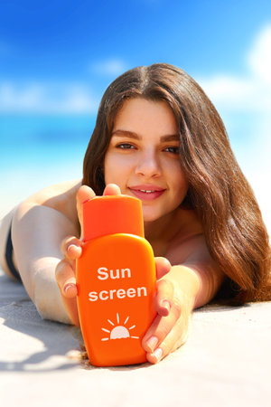 Young woman smiling on beach and holding sunscreen bottle in her hands