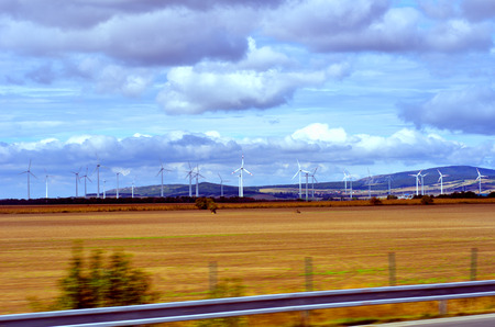 Blurred view on nature, cloudy sky, yellow field, wind power generators from window of moving car
