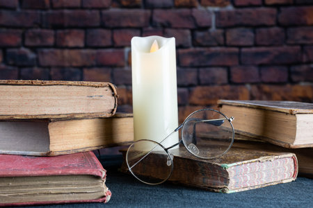 Glasses on stack of old books and candle on brick wall backgroundの写真素材
