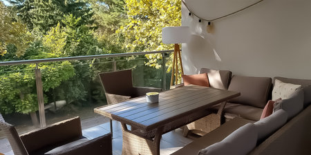 Summer scene of a spacious, comfortable loggia featuring a large soft sofa, two armchairs, a dining table, and stylish lamps. Surrounded by lush green trees and bushes, the airy setting radiates relaxation and eleganceの写真素材