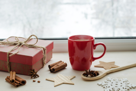 red cup with black strong espresso coffee with elevated steam on winter window sill, gift in beautiful box, anise stars, cinnamon sticks, wooden decorative elements, christmas time conceptの写真素材
