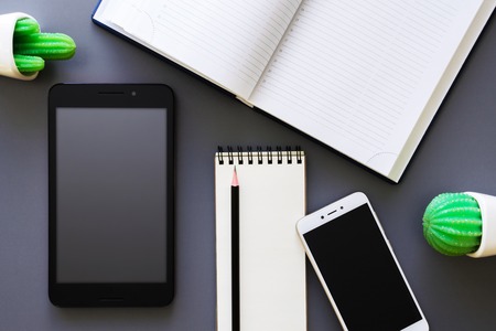 office working table desk with opened organizer, blank notepad, smartphone, tablet, pencil and green potted plant, dark background mock up, top viewの写真素材