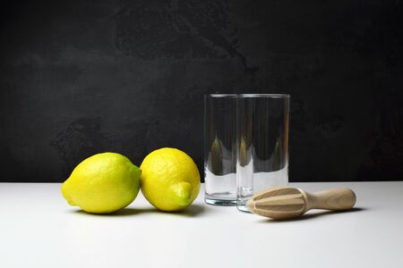 Two juicy lemons with wooden hand squeezer and two empty glasses on white table background over black backgdropの写真素材