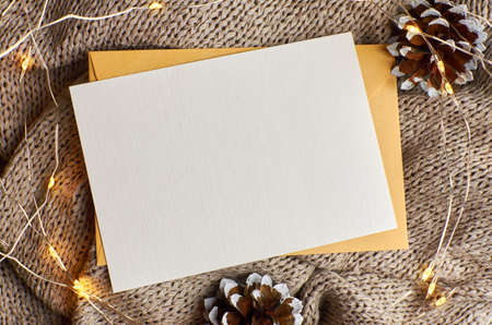 Greeting card mockup with garland and pine tree cones on knitted background. Christmas flat lay with copy space.の写真素材