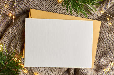 Greeting card mockup with garland and pine tree branches on knitted background. Christmas flat lay with copy space.の写真素材