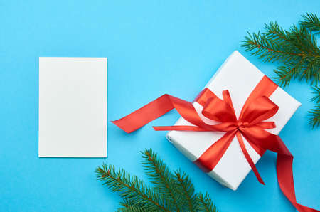 Greeting card mockup. Gift box with red ribbon with bow and fir tree branches on blue paper background. Winter holidays mockup, top view, copy space.の写真素材
