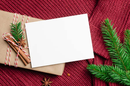 Christmas greeting card mockup with decorated gift box and fir tree branch on red knitted background