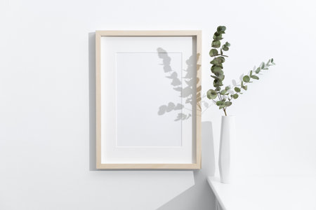 Vertical wooden frame mockup on white wall next to a modern vase with dried eucalyptus branches, set in a minimal and bright interior spaceの写真素材