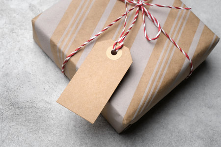 Kraft paper wrapped gift box tied with red and white twine, featuring a blank kraft tag on a light concrete surface, perfect for festive packaging mockupの写真素材