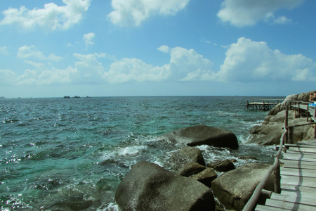 In the foreground there are large gray boulders and an old wooden bridge that goes into the sea. Dark green water, sea foam. Blue sky and clouds. Summer sunny day. Nobody here.の写真素材