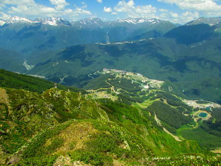 Mountain range on a Sunny summer day. In the foreground is a small hill covered with green grass and small white flowers. Downstairs overlooks the outdoor swimming pool. In the background a mountain range, the peaks of the snow. blue sky, clouds.の写真素材