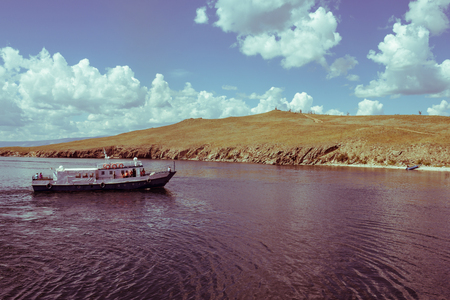 A small ship approaches the deserted shore. Calm blue water, rocky shore covered with yellow grass. A path leads to the top of the hill. At the stern of the ship is a crew, looking at the shore. Autumn sunny day. September.の写真素材