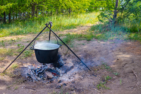 On the fire in the kettle cook soup. From the food there is steam. Sunny summer day. Around the fire trees and grass.の写真素材