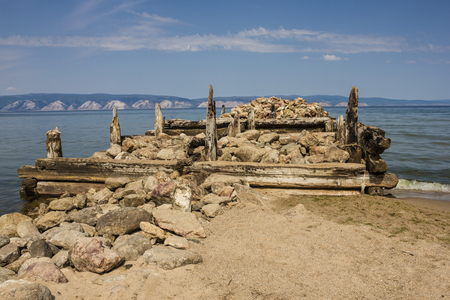 The ruined old pier goes into the distance. In the foreground are stones, logs, sand. Nobody here. On the opposite shore white mountains. Sunny day. Blue sky, small clouds.の写真素材