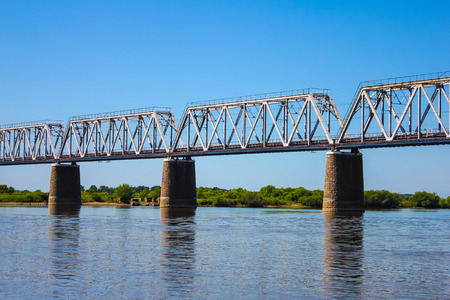 Bridge over the river close up. Bridge supports and spans. In the distance a sandy beach and green trees. The blue cloudless sky. Summer. Evening.の写真素材