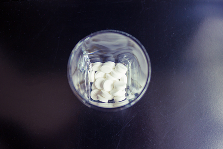 In a transparent glass are white tablets. Top view, daylight. Dark background. Medication, appointment of a doctor, drug therapy.の写真素材