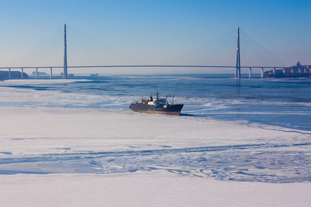 Seascape in blue tones. On the water a small ship, the sea partially frozen, covered with white snow. In the distance a huge cable-stayed bridge. Lateral solar illumination.の写真素材