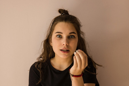A girl with wide eyes trying to say something. A modern teenager with long hair. The hand is raised to the mouth. Amazement, surprise. Light background.の写真素材