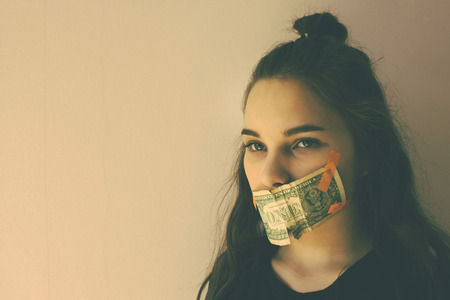 The girl's mouth is glued to the dollar. Forced silence, bribery, unwanted witness. Green eyes, long hair. Muted tones, copy space.の写真素材