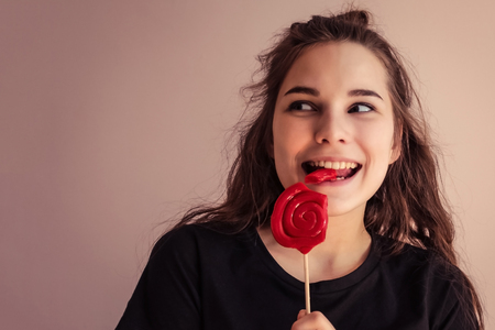 A smiling girl is eating a big red candy on a stick. Startling smile, looking away, long hair, black T-shirt. Light background, copy space, vignetting.の写真素材