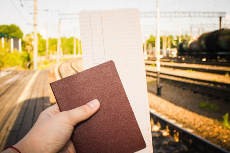 A hand holds a railway ticket and a passport. In the background there is a rails, a railway station. Anticipation of travel, voyage, long road. Sunlight, summer day.の写真素材