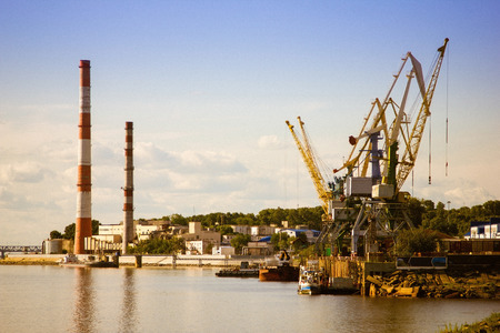 Industrial city, river port. Large pipes, loading cranes, ships. Brown water, blue sky. Far away on the hill of the house. Sunny summer evening.の写真素材