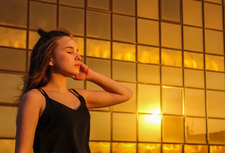 Modern girl, teenager, against a glass wall background. The windows reflect the yellow sun, sunset. Urban youth, a new generation.の写真素材