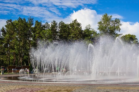 Bright sunny day, heat. Large city fountain, high streams of water, white splashes. In the distance are tall green trees, benches.の写真素材