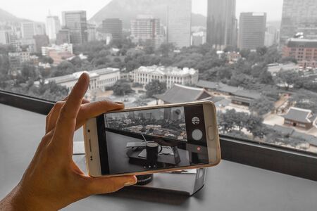 A cup of morning coffee on the background of Seoul. Modern buildings mixed with old houses, downtown. Female hand takes a photo on a smartphone. Muted tones.の写真素材